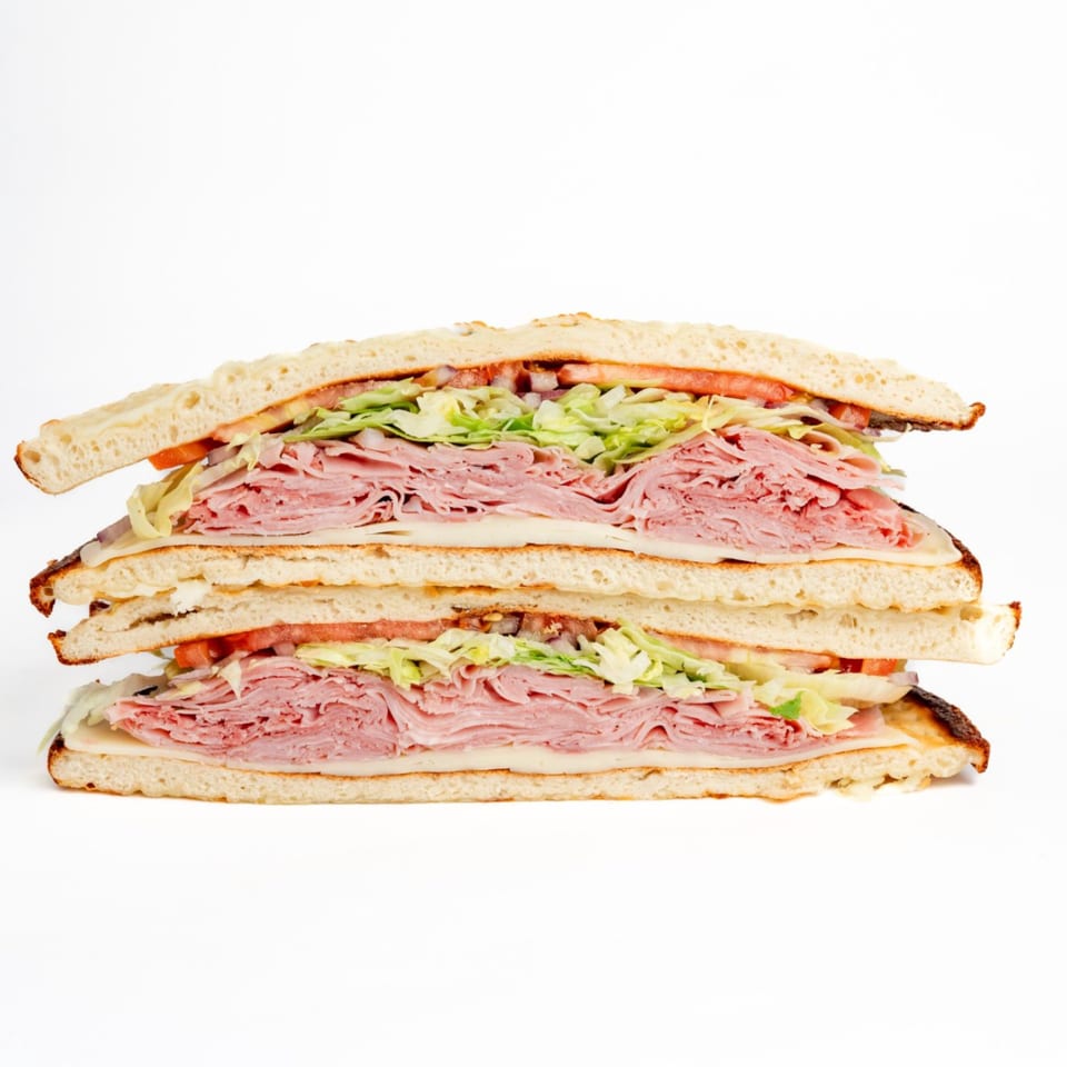 Two halves of a large deli sandwich, stacked, filled with ham, cheese, lettuce, and tomato.