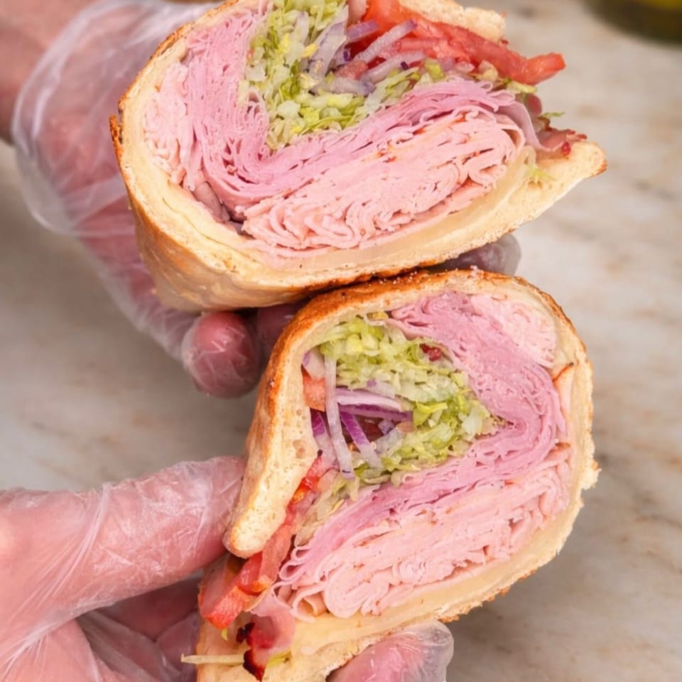Gloved hands holding a large deli sub sandwich, cut in half, revealing layers of meat, cheese, and vegetables.
